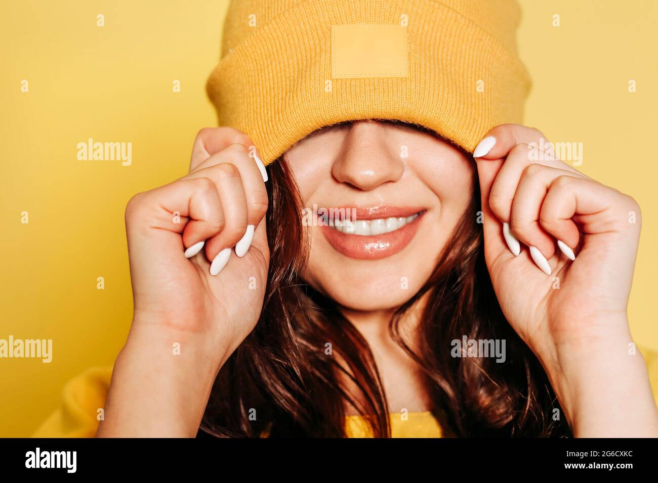 Fröhliche junge Hündin im trendigen gelben Sweatshirt und Strickmütze Die Augen vor gelbem Hintergrund lächeln hell Stockfoto