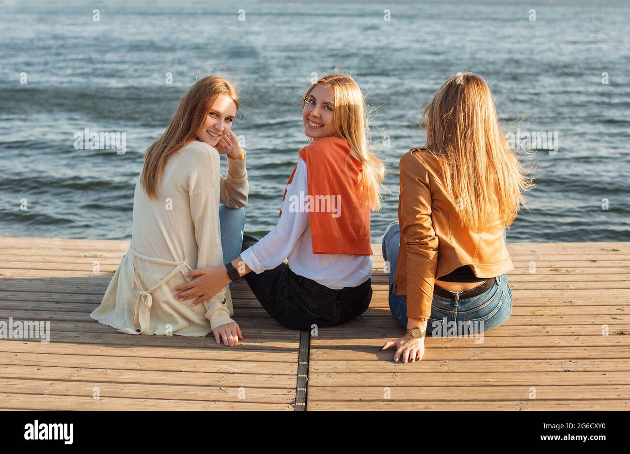 Rückansicht der fröhlichen jungen Frauen in lässiger Kleidung sitzen Auf dem Kai in der Nähe des Wassers und Blick auf die Kamera über Schultern Stockfoto