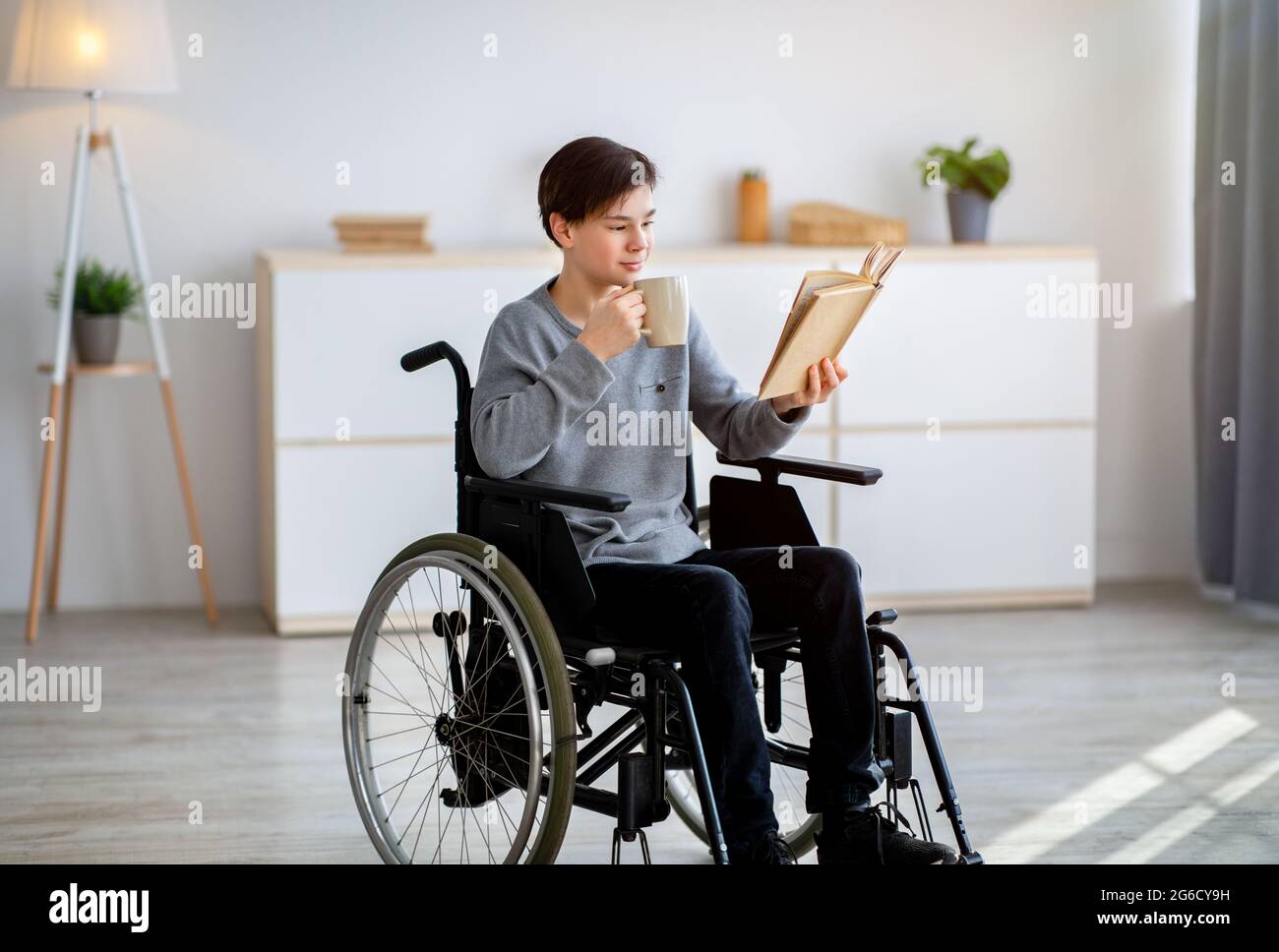 Positiver behinderter Teenager Junge im Rollstuhl, der Interessantes Buch liest und zu Hause Kaffee trinkt Stockfoto