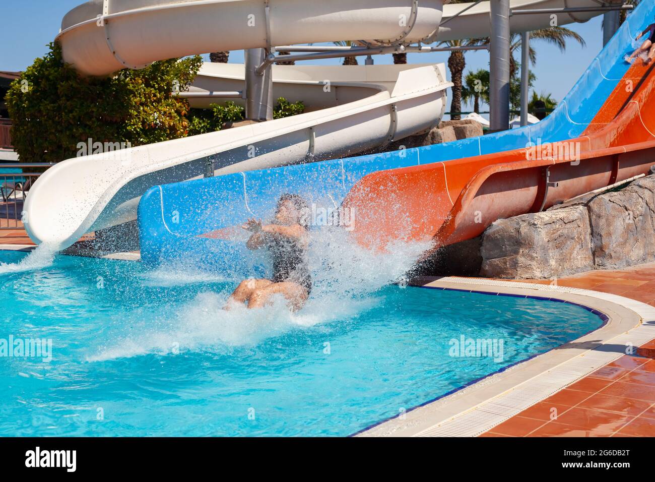 Fröhliches kleines Mädchen, das in den Sommerferien auf der Wasserrutsche untergeht und in das Schwimmbad mit großem Planschbecken im Wasserpark eindringt. Sommerurlaub für Kinder im Freien. Stockfoto