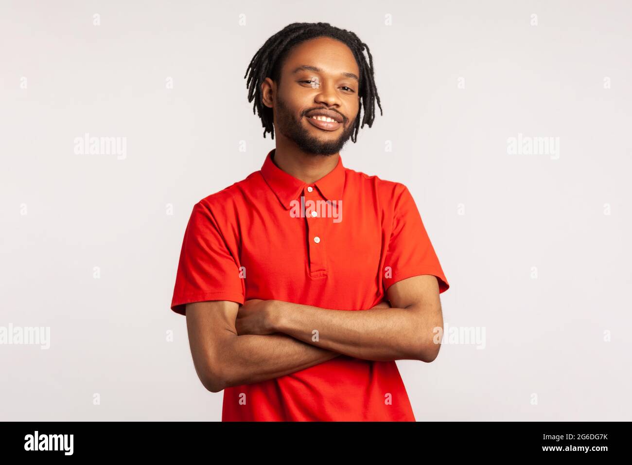 Junger erwachsener bärtiger Mann mit Dreadlocks in einem roten, lässigen T-Shirt, der mit selbstbewusster Gesichtsausdruck und gekreuzten Armen posiert und lächelnd auf die Kamera blickt Stockfoto