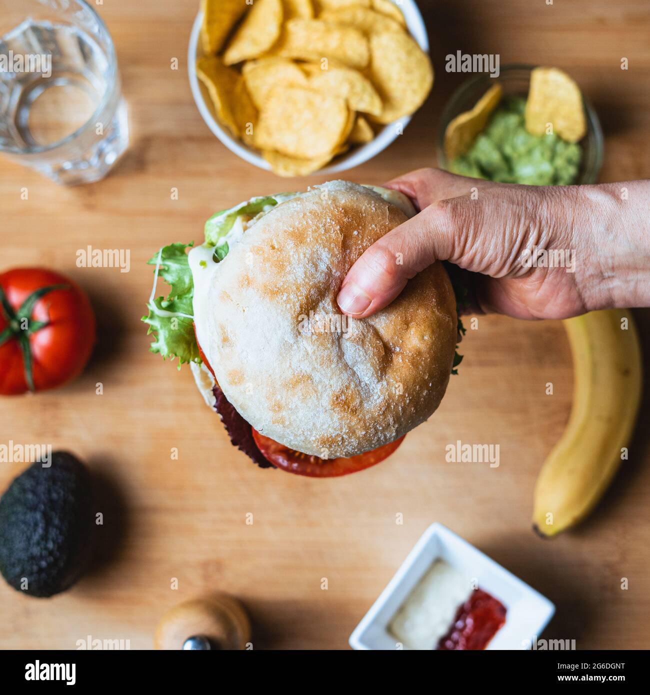 Frischer, leckerer Burger mit rustikalem Brötchen, umgeben von Nachos mit Guacamole zum Eintauchen, ein Wasserglas, eine Tomate, eine Avocado, Pfeffer, Ketchup und Mayonnaise Stockfoto
