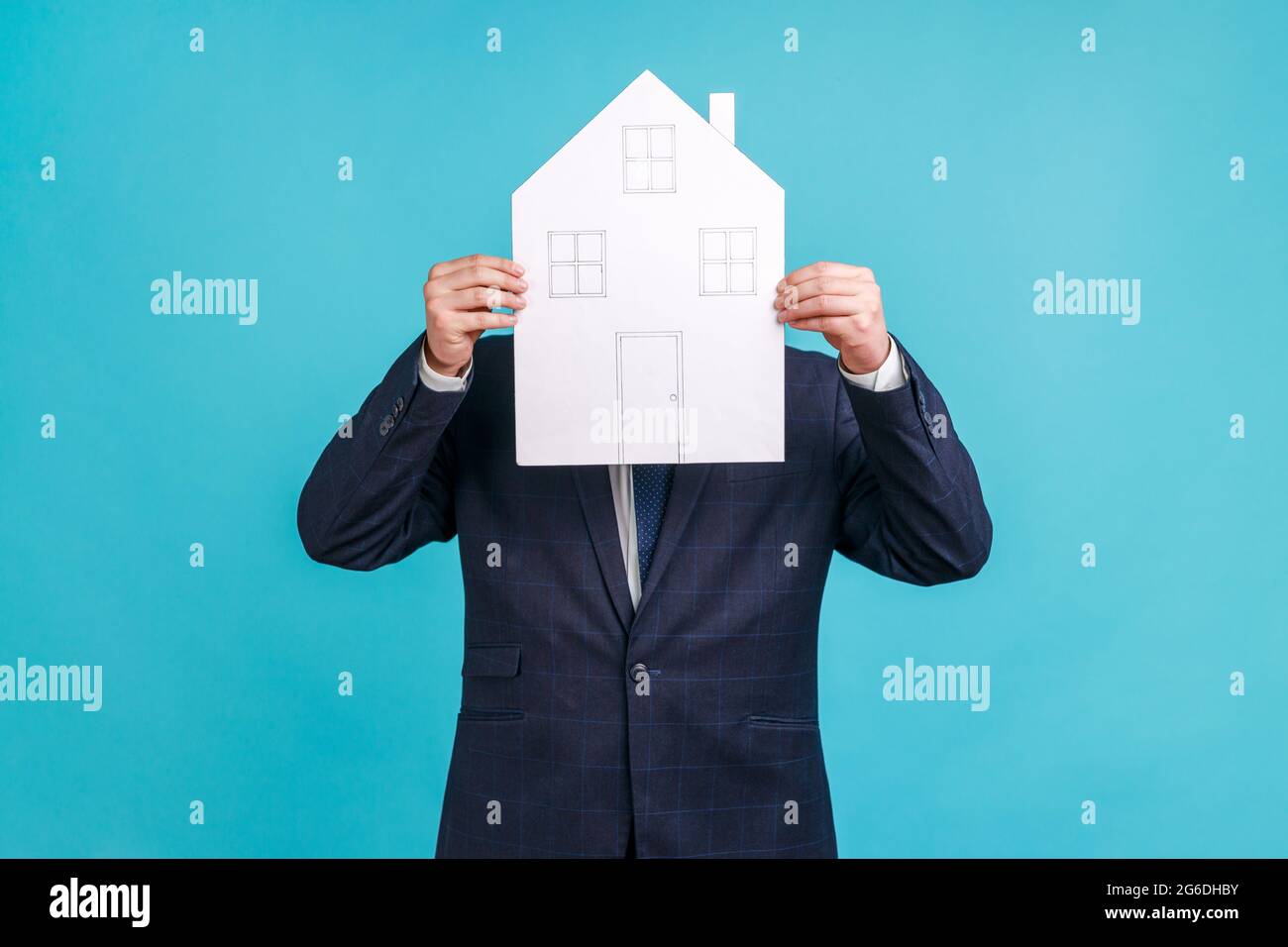 Unbekannter Mann in elegantem Anzug, der sein Gesicht hinter einem Papierhaus mit gezogenen Fenstern und Türen, einem staatlichen Wohnungsbauprogramm und einer Immobilienagentur versteckt. Indoo Stockfoto