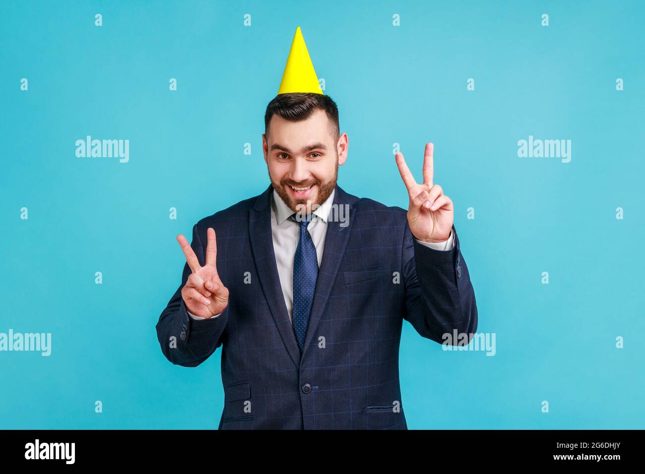 Glücklicher bärtiger Kerl mit witzigem Partykegel am Kopf, der mit beiden Händen ein Siegeszeichen zeigt, mit toothy Lächeln auf die Kamera schaut und Geburtstag holida feiert Stockfoto