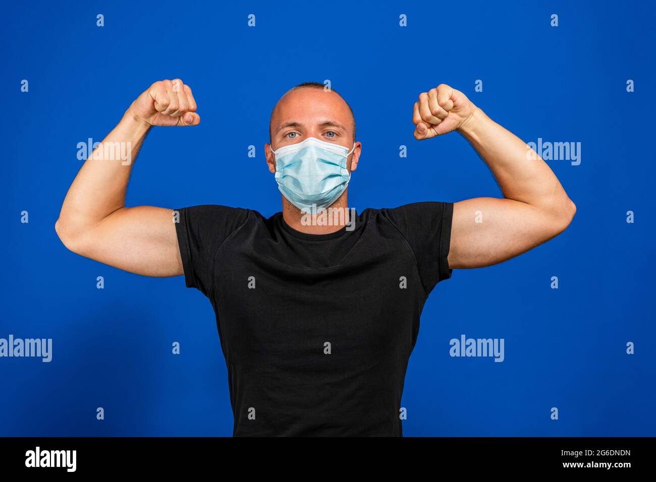 Junger gutaussehender Mann in medizinischer Maske und gelbem T-Shirt isoliert auf blauem Hintergrund. Starke Person zeigt Arm Muskel, zuversichtlich und stolz auf die Macht zu d Stockfoto
