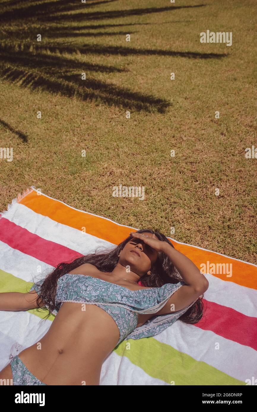 Vertikale Ansicht schwarze indische Womanliegend in einem Handtuch auf dem Gras mit Palmen Schatten auf ihrem Körper Sonnenbaden nach einem Bad im Pool. Summer Vibes 2021. Stockfoto