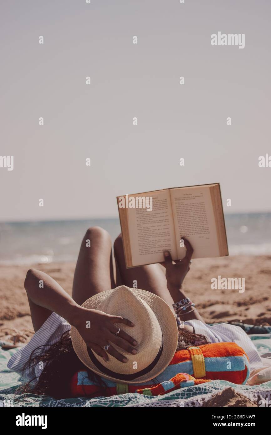 Vertikale Rückansicht schwarze inderin, die am Strand liegt und ein Buch liest, während sie ihren Sommerhut an einem sonnigen, windigen Sommertag hält. Sommerstimmung 2021 Stockfoto