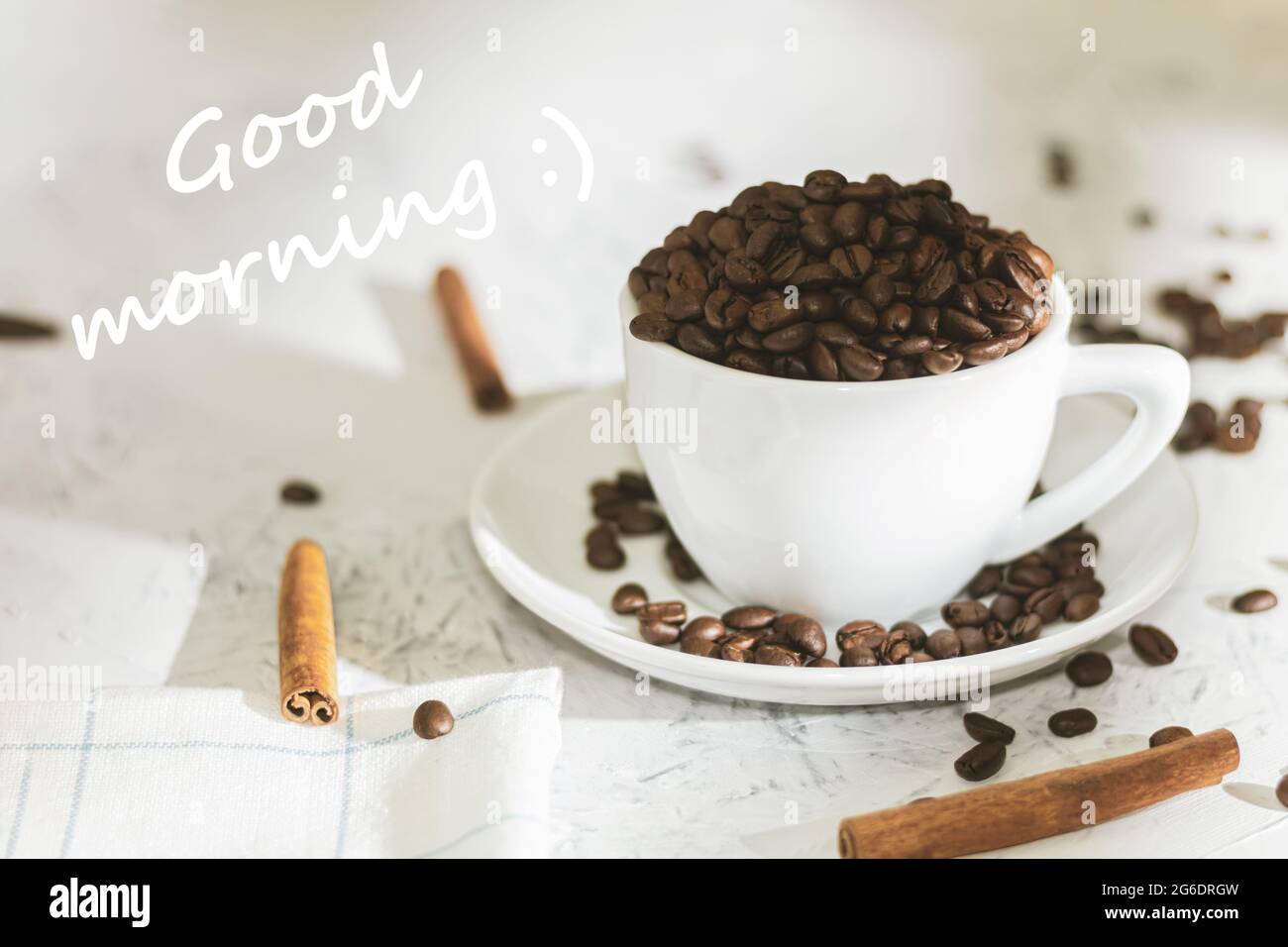 Weiße Tasse mit Kaffeebohnen und dem Text Guten Morgen. Guten Morgen geschrieben. Kaffeebohnen in einer Kaffeetasse und Zimtstangen. Hintergrund mit Text Stockfoto
