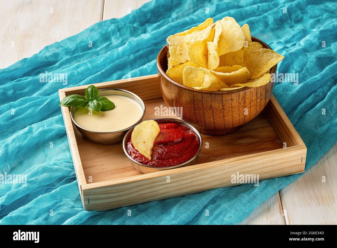 Leckere Kartoffelchips in einer Holzschüssel und kleine Metallschüsseln mit dickem Tomatenketchup und Mayonnaise auf einem rechteckigen Holztablett auf türkisfarbenem Tuch. Stockfoto