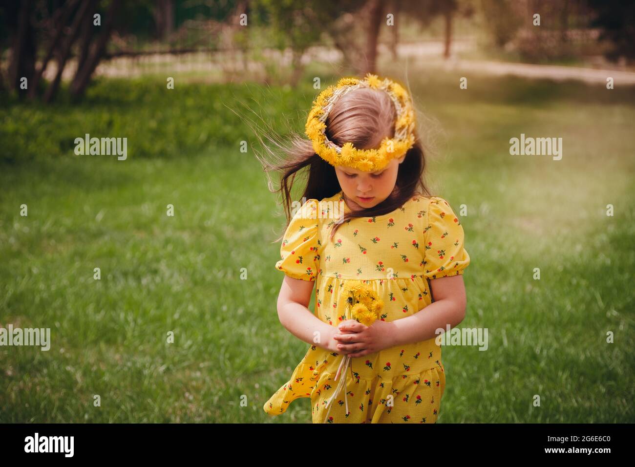 Ein süßes Mädchen mit lockeren Haaren und einem Kranz aus gelben Delikten auf ihrem Kopf sieht einen Strauß von Delikten an, wobei ihr Kopf in einem schönen Grün abgesenkt ist Stockfoto