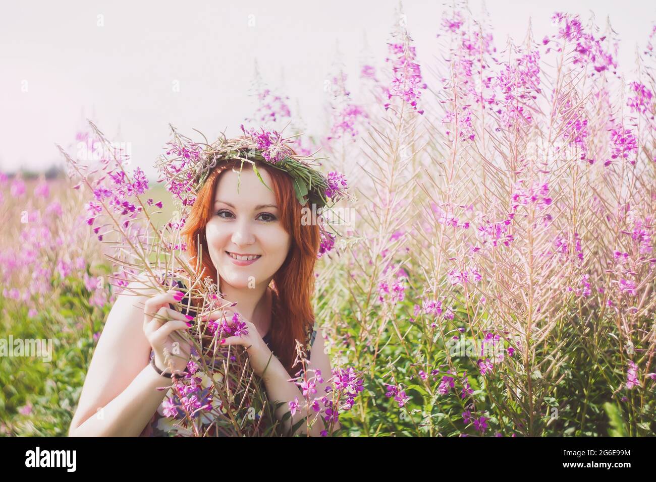 Porträt einer jungen, schönen rothaarigen Frau mit einem Kranz auf ihrem Kopf. Lächelnde Dame steht auf einem Feld mit wilden rosa Blumen. Stockfoto