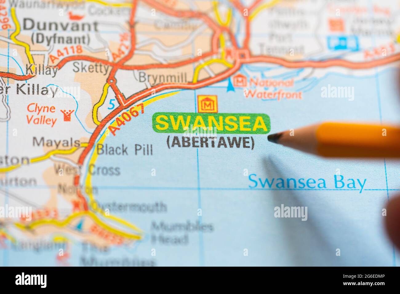Eine Makroaufnahme einer Seite in einem gedruckten Roadmap-Atlas mit einer Männerhand, die einen Bleistift hält und auf die Stadt Swansea in Wales zeigt Stockfoto