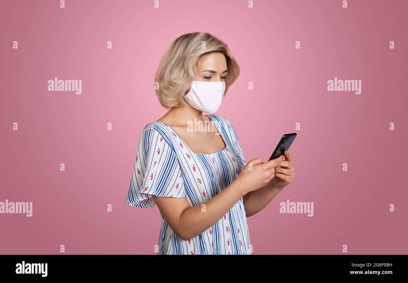 Die kaukasische Frau plaudert auf dem Handy, während sie eine medizinische Maske an einer roten Studiowand trägt Stockfoto