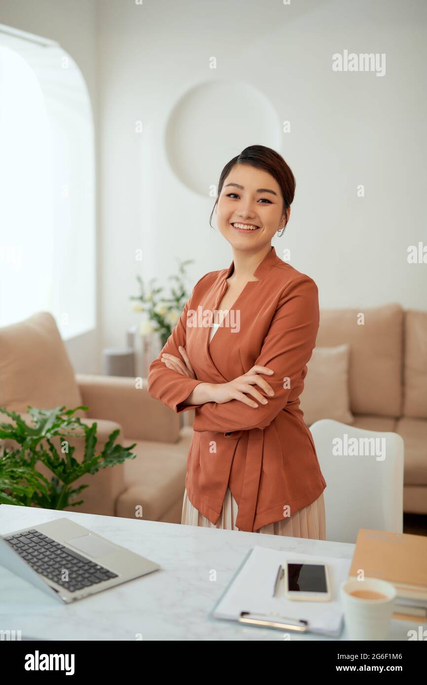 Lächelnde Frau mit verschränkten Armen steht an ihrem Arbeitsplatz im Home Office Stockfoto
