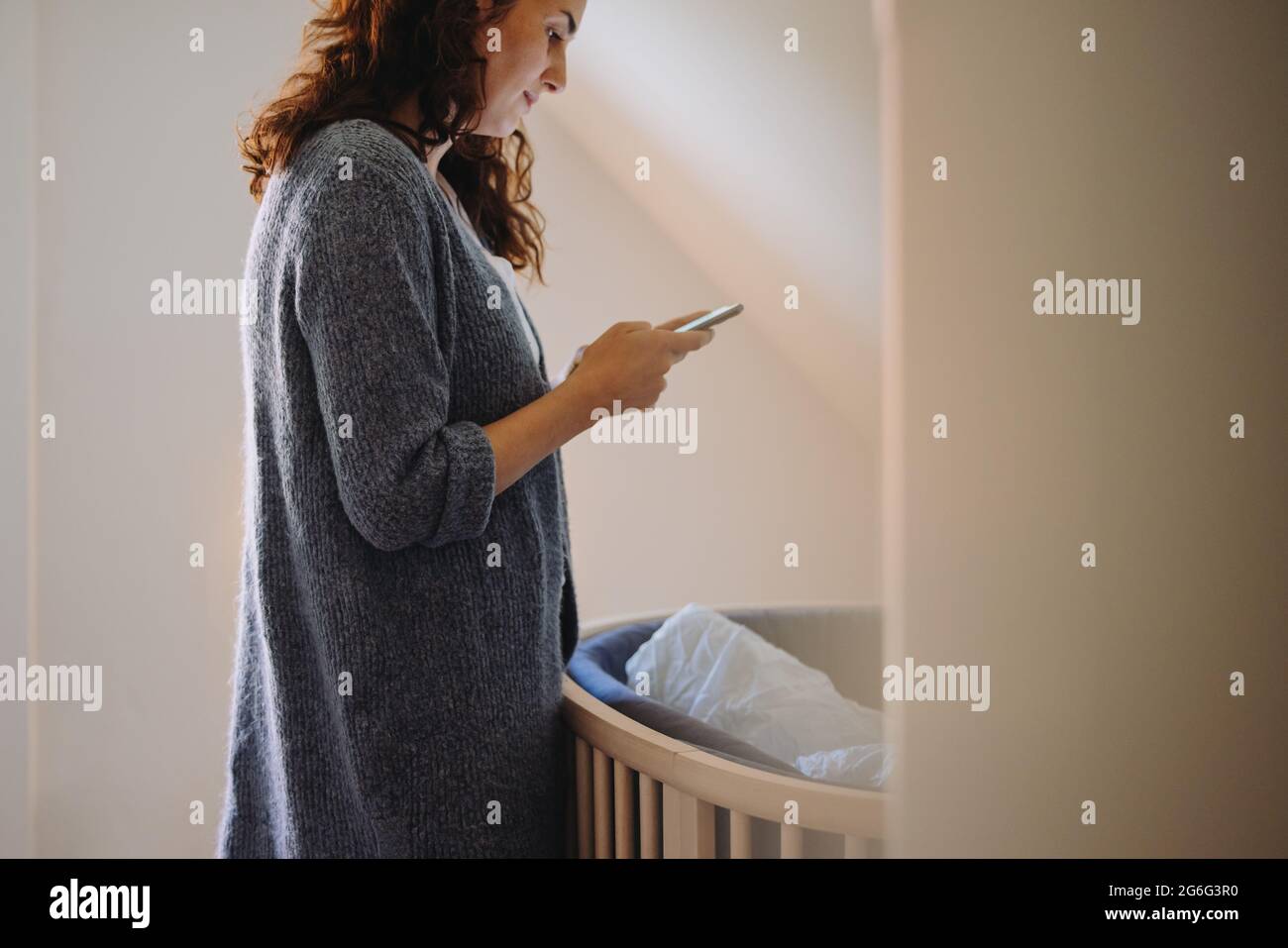 Weibliche SMS auf ihrem Handy zu Hause. Mutter, die mit dem Mobiltelefon neben einem Kinderbett steht. Stockfoto