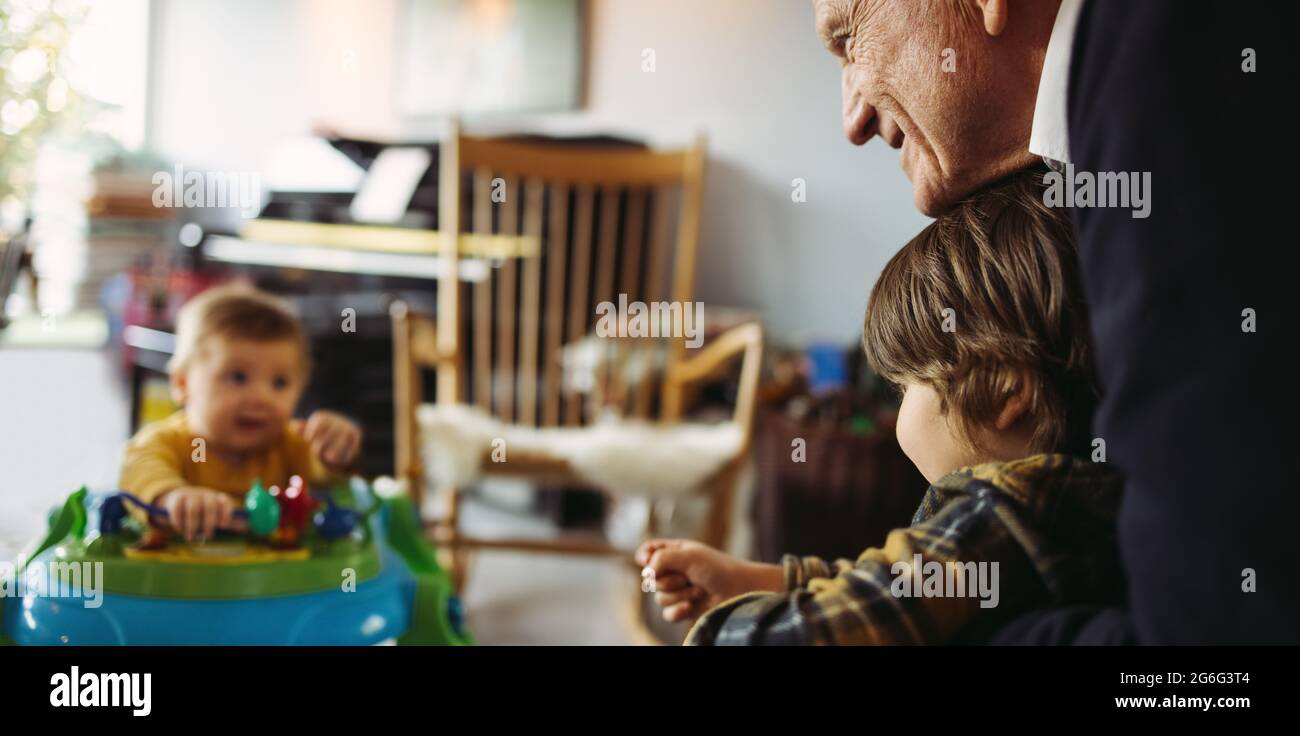 Älterer Mann hält seinen Enkel und sieht das Baby zu Hause auf einem Spaziergänger sitzen. Opa mit Enkeln zu Hause. Stockfoto