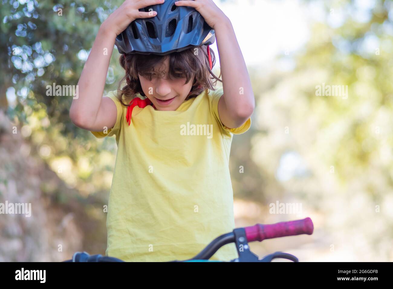 Kleiner lächelnder Junge mit langen Haaren und gelbem T-Shirt, der seinen Schutzhelm justiert, während er mit dem Fahrrad durch die Straßen seiner Stadt fährt Stockfoto