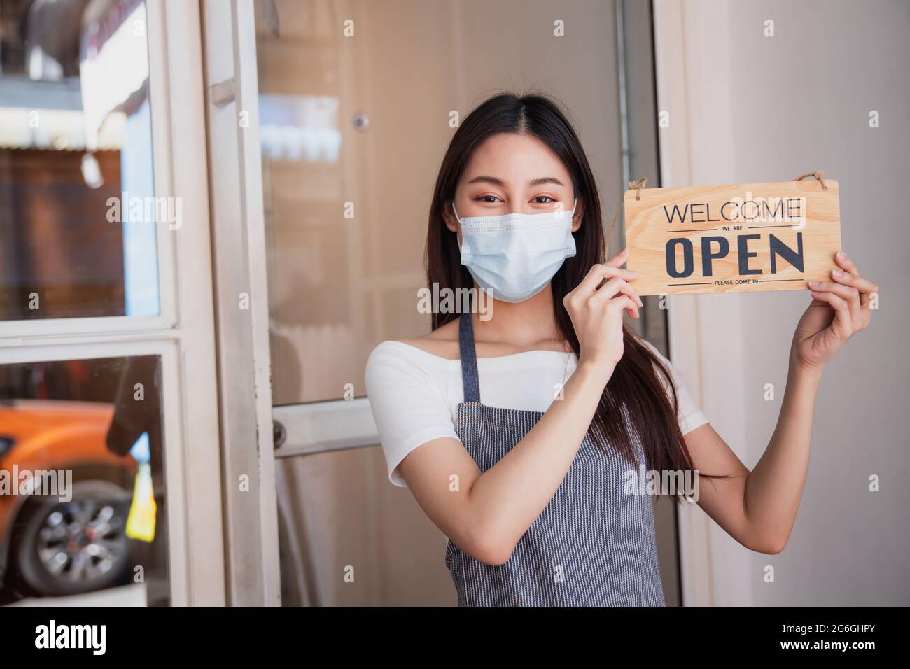 Die Inhaberin des asiatischen Cafés trägt eine Maske, die Frau hält ein Schild, das nach der Stadtsperre geöffnet wird, und ist bereit, zu verkaufen und zu arbeiten Stockfoto