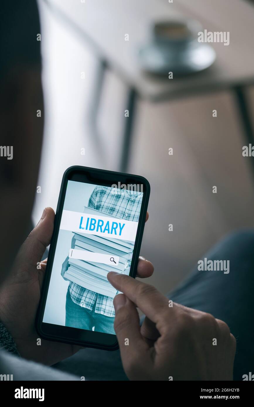 Ein junger Mann, der an einem Couchtisch in seinem Wohnzimmer sitzt, sucht mit seinem Smartphone ein eBook, das er mit einer simulierten SE auf einer Online-Bibliothek ausleihen kann Stockfoto