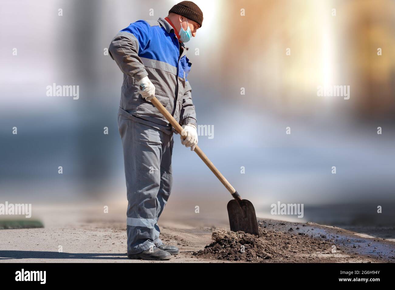 Ein Arbeiter in einer medizinischen Maske und Arbeitskleidung steht mit einer Schaufel auf einem verschwommenen Hintergrund. Körperliche Arbeit. Arbeitslosigkeit während der Pandemie. Beschäftigung und Arbeit. Stockfoto
