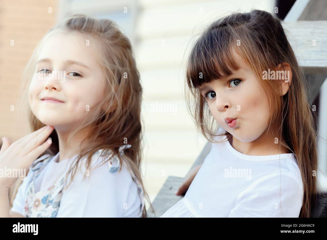 Zwei schöne Mädchen Freundinnen sitzen an einem Sommertag auf einer Holztreppe und blicken in die Kamera und zappeln. Kinderportrait. Echte Emotionen. Stockfoto
