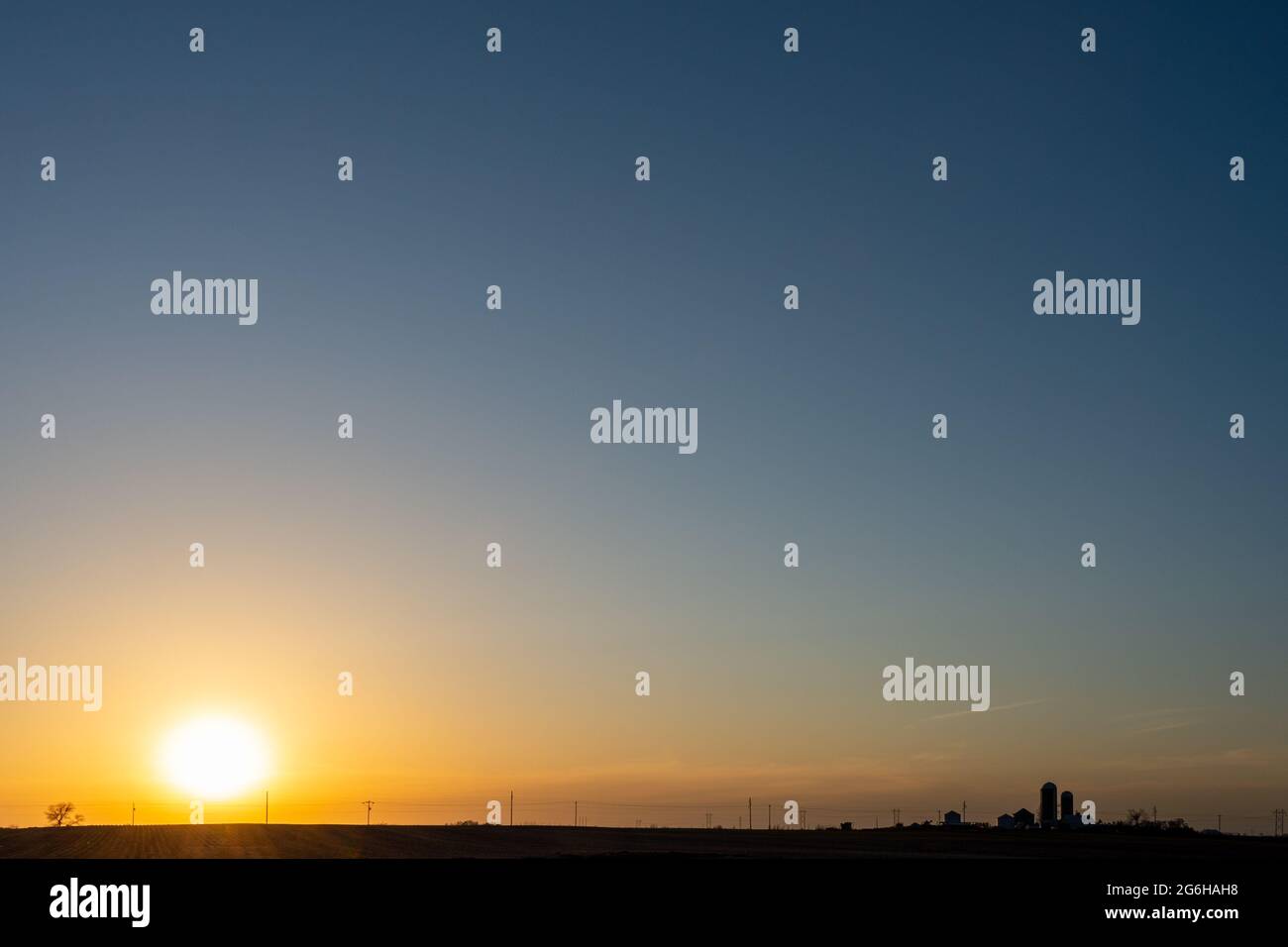 Eine untergehende Sonne umrisset den Horizont und entfernte Bauernhäuser und Silos. Stockfoto