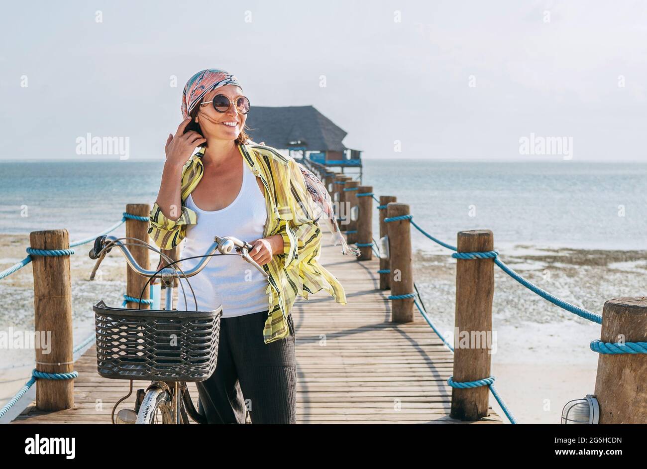 Porträt einer lächelnden Frau in leichte Sommerkleidung gekleidet, Sonnenbrille mit Fahrrad auf dem hölzernen Seebrücke am Sandstrand von Sansibar.sorgloser Urlaub Stockfoto