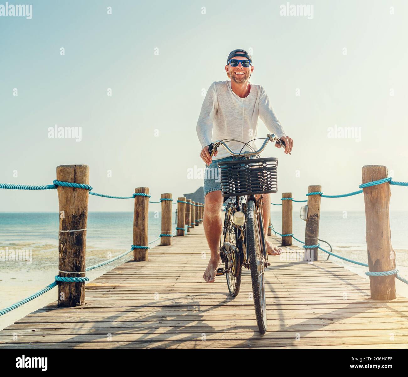 Porträt eines glücklichen lächelnden Mannes, gekleidet in leichte Sommerkleidung und Sonnenbrille, der auf dem hölzernen Seebrücke mit dem Fahrrad fährt. Sorgloser Urlaub im tropischen Stockfoto
