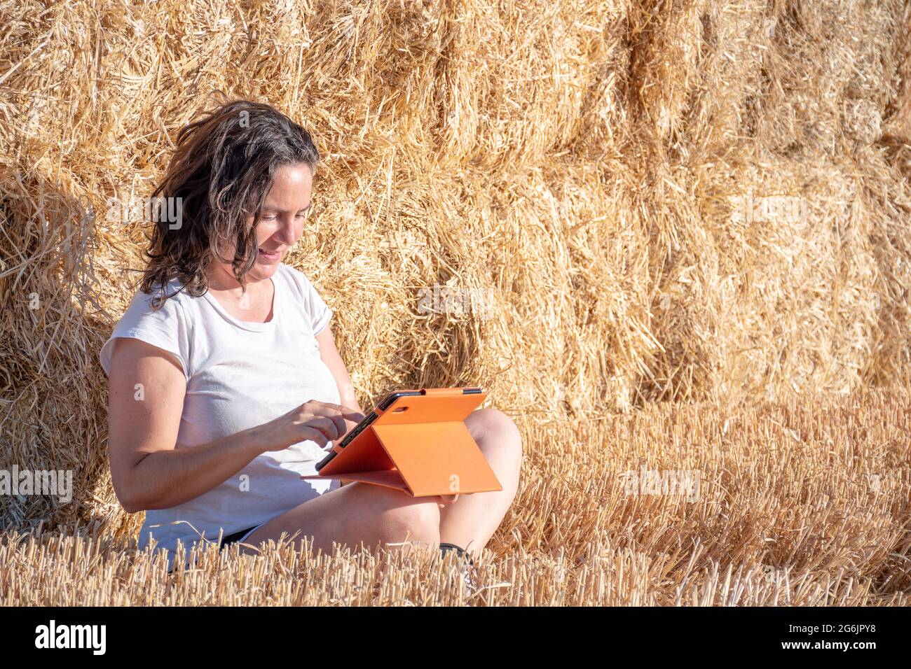 Die brünette Latina-Frau mittleren Alters sitzt auf dem Boden auf einem Feld und lehnt sich mit einer Tablette mit orangefarbenem Deckel auf einen Haufen Strohballen. Technologisch und Stockfoto