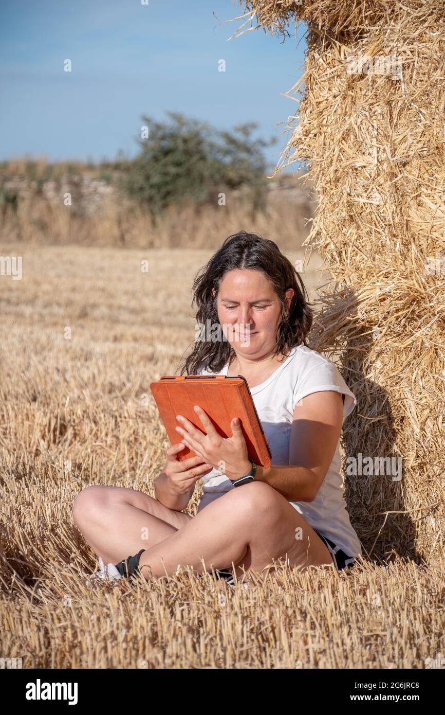 Die brünette Latina-Frau mittleren Alters sitzt auf dem Boden auf einem Feld und lehnt sich mit einer Tablette mit orangefarbenem Deckel auf einen Haufen Strohballen. Technologisch und Stockfoto