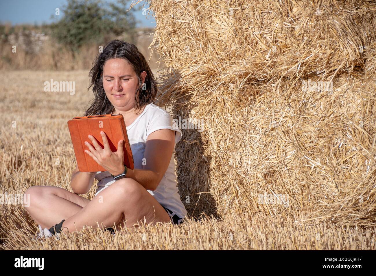 Die brünette Latina-Frau mittleren Alters sitzt auf dem Boden auf einem Feld und lehnt sich mit einer Tablette mit orangefarbenem Deckel auf einen Haufen Strohballen. Technologisch und Stockfoto