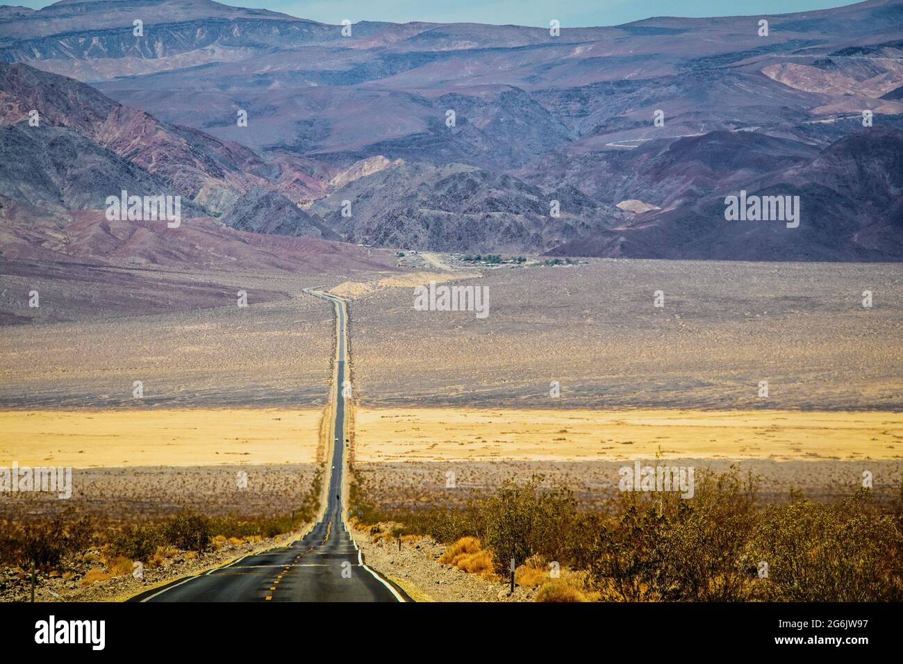 Zweispurige Blacktop-Autobahn, die in der Hitze schwankt, die sich in die fernen Berge ausdehnt, mit einem Sandband, das die raue strukturierte Sageburst-Terr durchschneidet Stockfoto