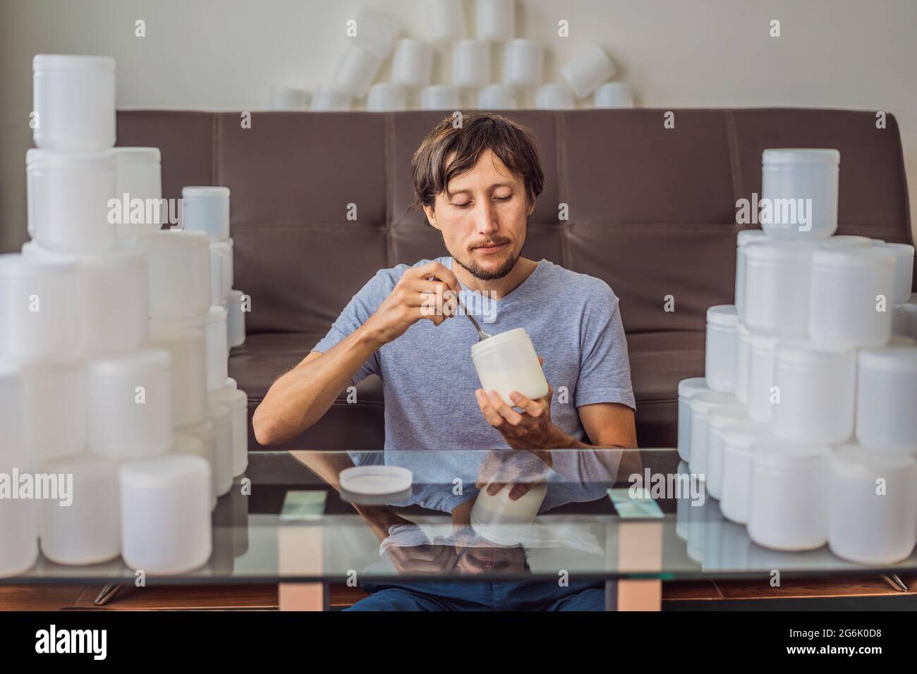 Der Mann isst Joghurt und ist von vielen Joghurtgläsern umgeben, die er in einem Jahr verwendet. Eine riesige Menge von Plastikbehältern für Lebensmittel. Übermäßiger Verbrauch von Stockfoto