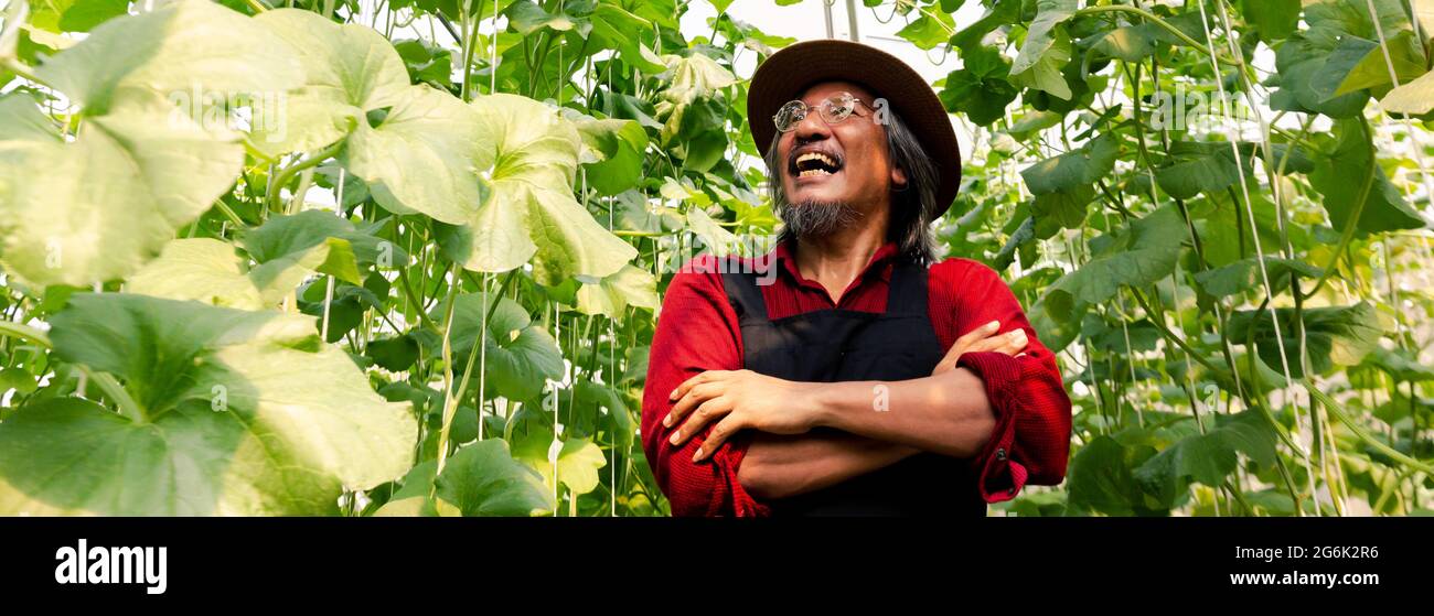 Älterer Bauer mittleren Alters mit den Armen mit glücklich gekreuzt Ein Lächeln mit Strohhut in roter Farmuniform Im Sommer im Garten des Bauernhofs Stockfoto