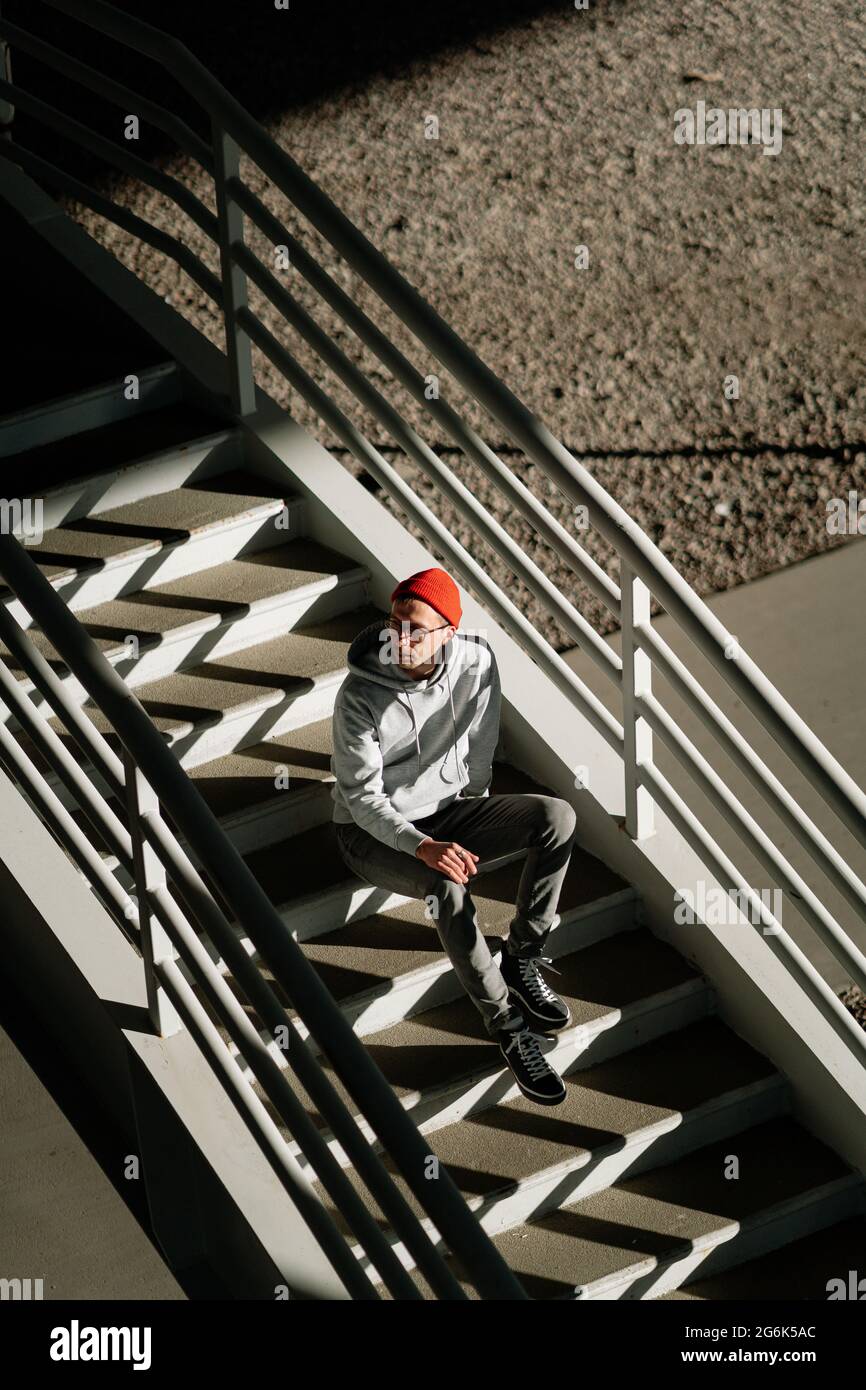 Einsamer junger Mann in Brille sitzt allein auf Betontreppen im städtischen Gebäude oder Stadtraum, oberhalb der Ansicht Stockfoto