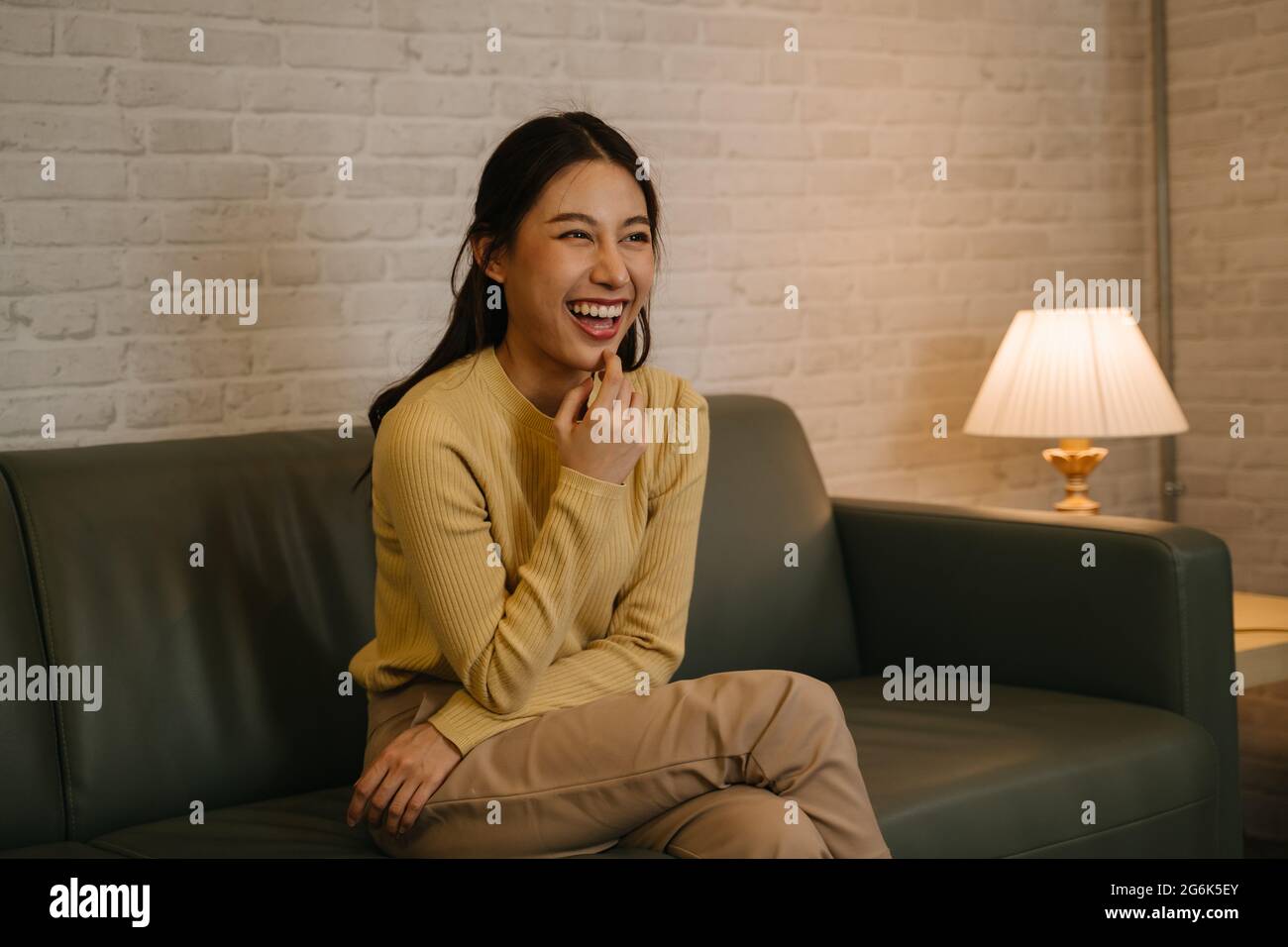 Fröhliche junge asiatische Frau, die zu Hause auf der Couch sitzt und lacht, entspannt, während sie abends Comedy-Shows und Filme im Fernsehen sieht Stockfoto