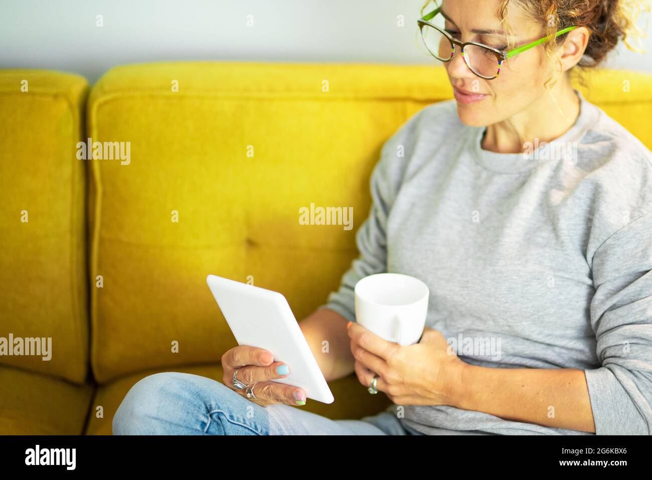 Nahaufnahme Porträt einer reifen hübschen Frau, die ein Buch auf einem Tablet liest, mit einer Brille, die zu Hause auf der gelben Couch sitzt, in der Pause Freizeit-Aktivität Stockfoto