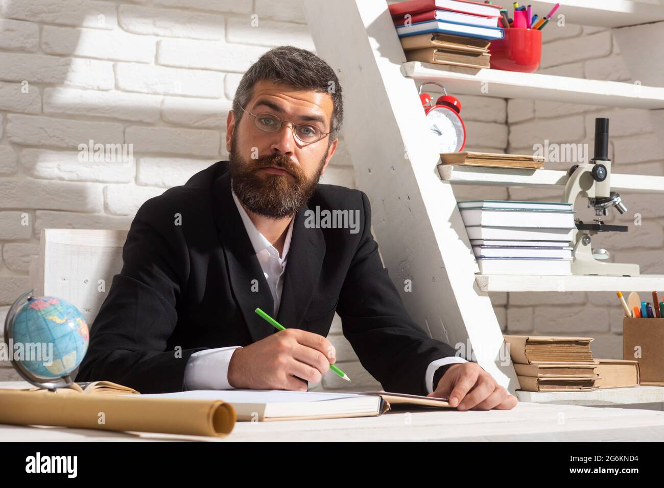 Porträt eines denkenden männlichen Lehrers im Klassenzimmer. Ernsthafter Lehrer Mann sitzt auf dem Schreibtisch und schreibt Text im Buch im Klassenzimmer. Stockfoto