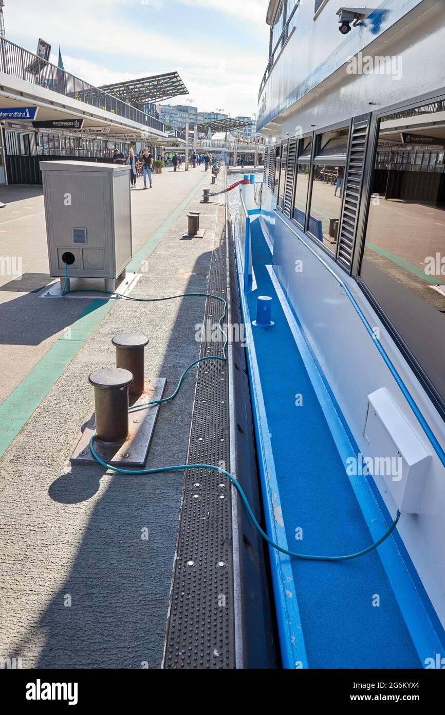 07. Juli 2021, Hamburg: Ein Stromkabel führt von einem Stromkasten (l) auf dem Ponton der Landungsbrücke 4 zu einem Schiff. Mit der heutigen Einweihung des Landkraftwerks an den Hamburg Landungsbrücken wurde die erste der geplanten Landkraftanlagen für die Binnenschifffahrt im Hamburger Hafen eröffnet. Das Landstromsystem wird es Fahrgastschiffen ermöglichen, sich im Hafen mit Strom aus erneuerbaren Energiequellen zu versorgen und ihre Dieselgeneratoren an Bord für die Stromerzeugung abzuschalten. Dies reduziert Luftschadstoffe wie Stickstoff und Schwefeloxide, klimaschädliches CO2 Stockfoto