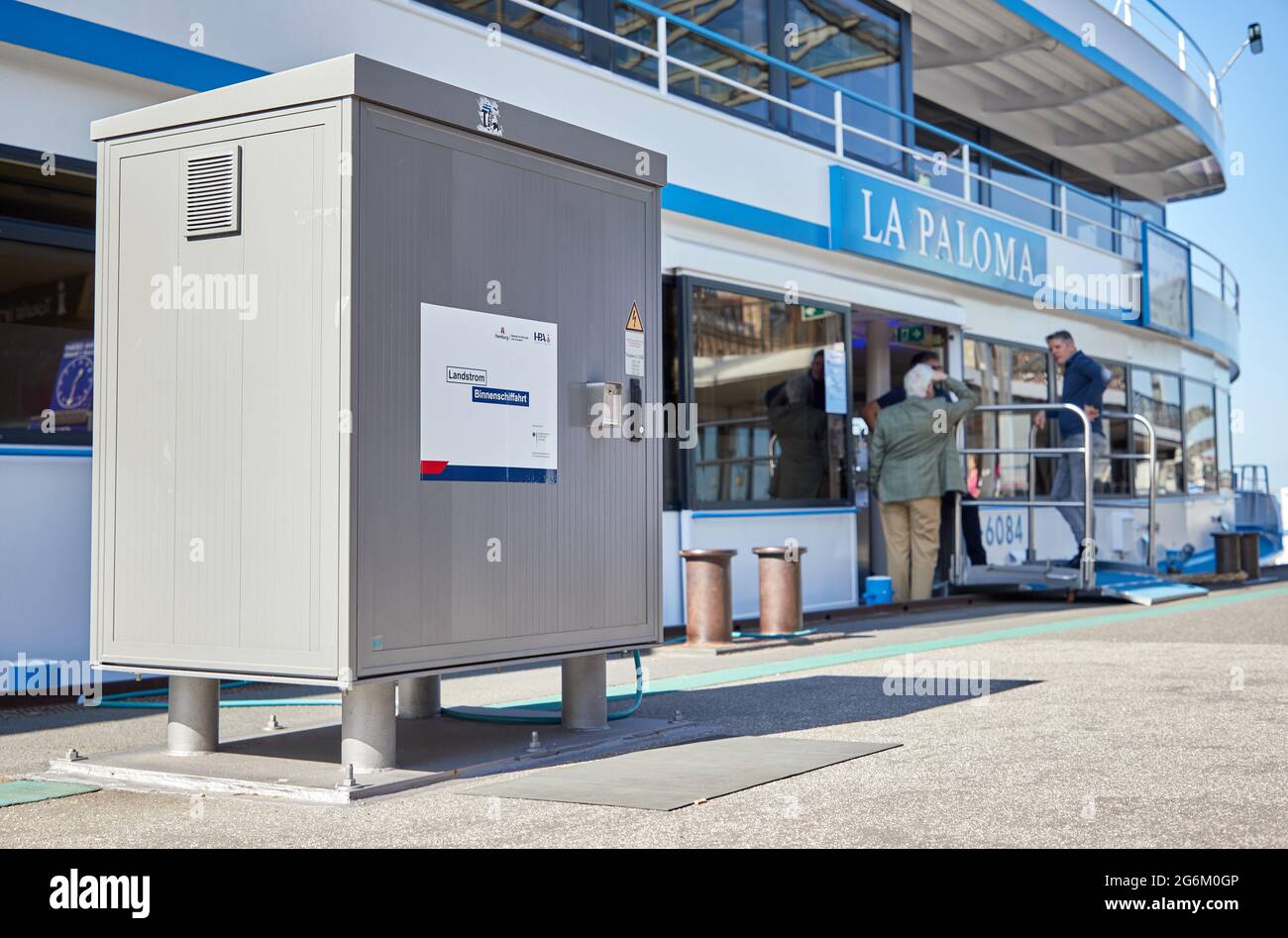 07. Juli 2021, Hamburg: Ein Stromkabel führt von einem Stromkasten (l) auf dem Ponton der Landungsbrücke 4 zum Hafenkreuzfahrtschiff La Paloma. Mit der heutigen Einweihung des Landkraftwerks an den Hamburg Landungsbrücken wurde die erste der geplanten Landkraftanlagen für die Binnenschifffahrt im Hamburger Hafen eröffnet. Das Landstromsystem wird es den Fahrgastschiffen ermöglichen, sich im Hafen mit Strom aus erneuerbaren Energien zu versorgen und ihre Dieselgeneratoren an Bord zur Stromerzeugung abzuschalten. Dies reduziert Luftschadstoffe wie Stickstoff und Schwefel oxid Stockfoto