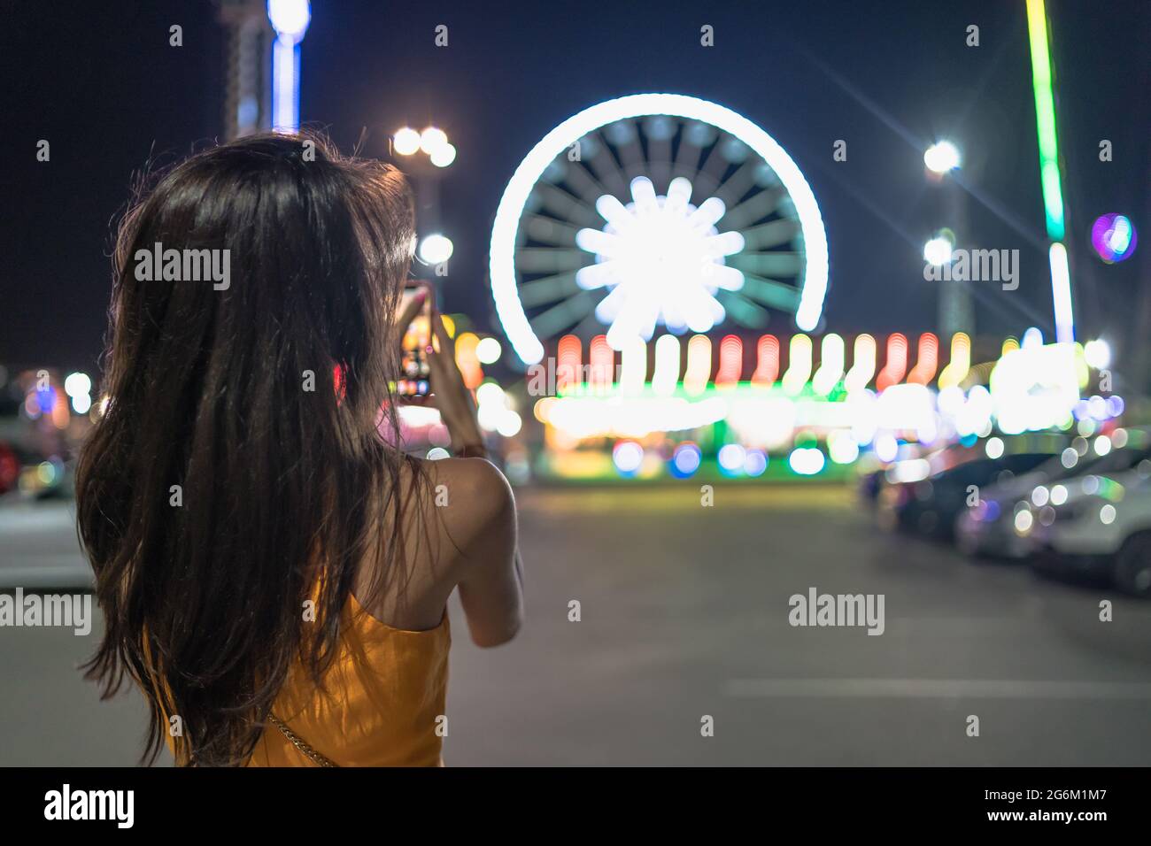 Porträt einer jungen Frau, die in der Nacht das beleuchtete Riesenrad im Vergnügungspark fotografiert Stockfoto