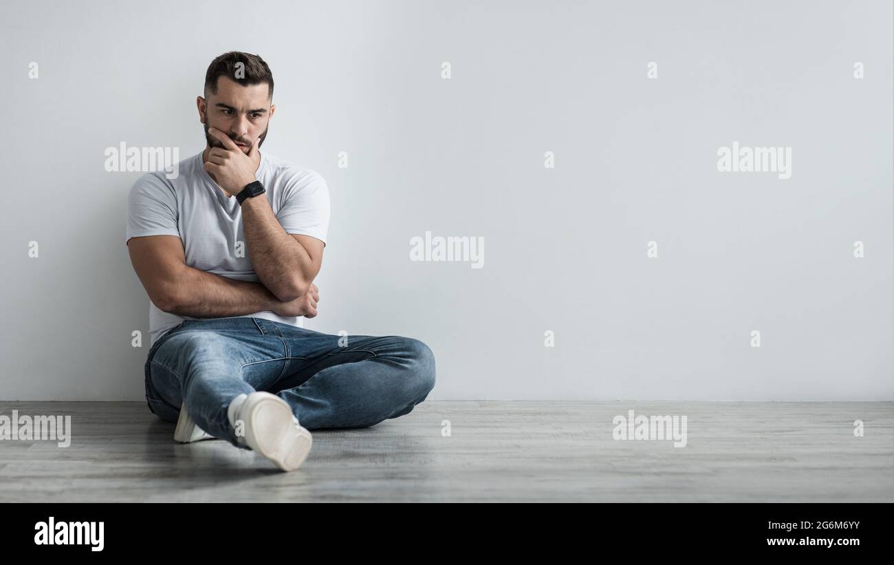 Ein junger depressiver Mann sitzt zu Hause an einer grauen Wand und fühlt sich unglücklich, einsam und traurig in Depressionen Stockfoto