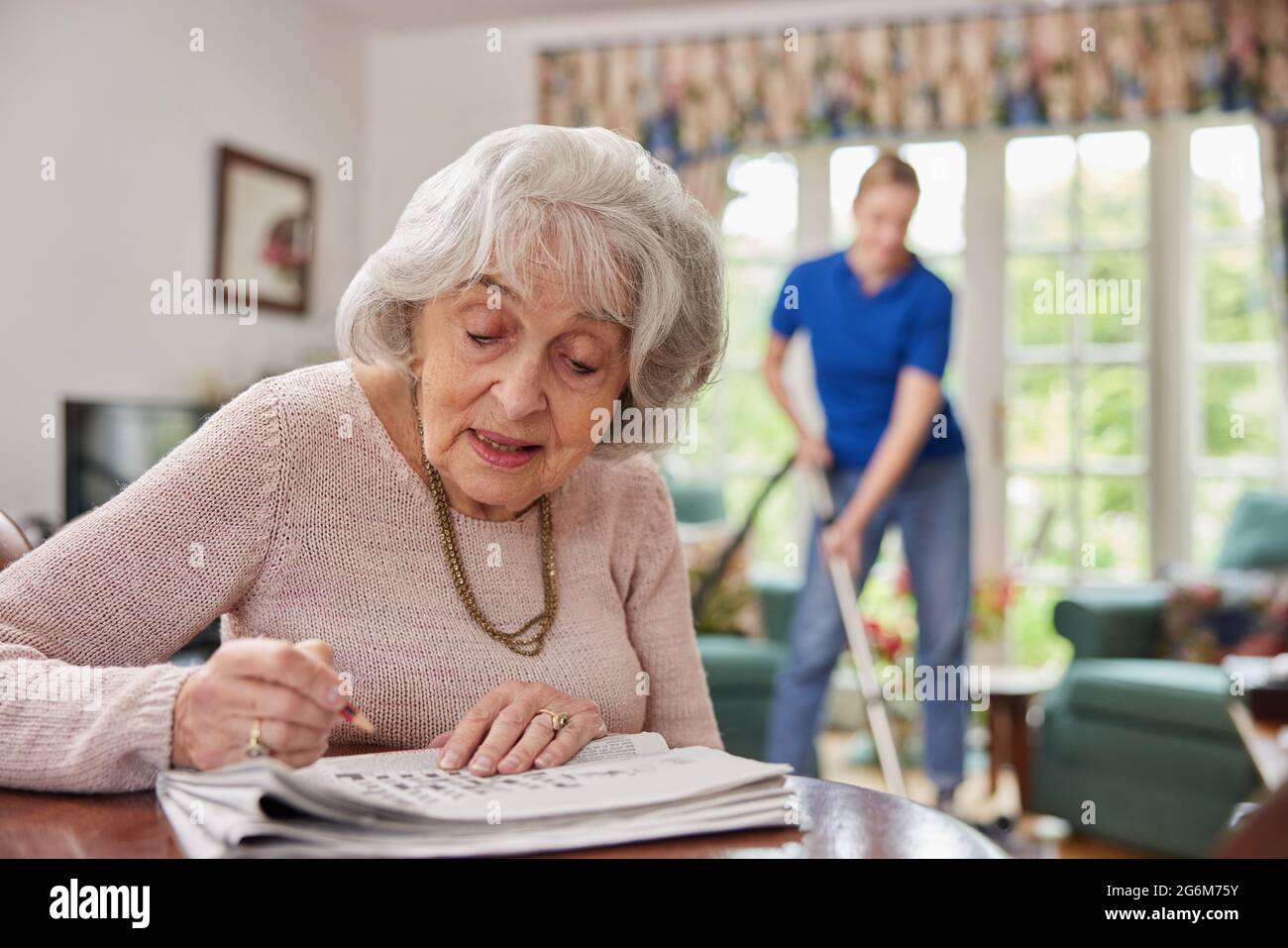 Weibliche Heim Hilfe Reinigung Haus Mit Staubsauger, Während Ältere Frau Tut Kreuzworträtsel In Der Zeitung Stockfoto
