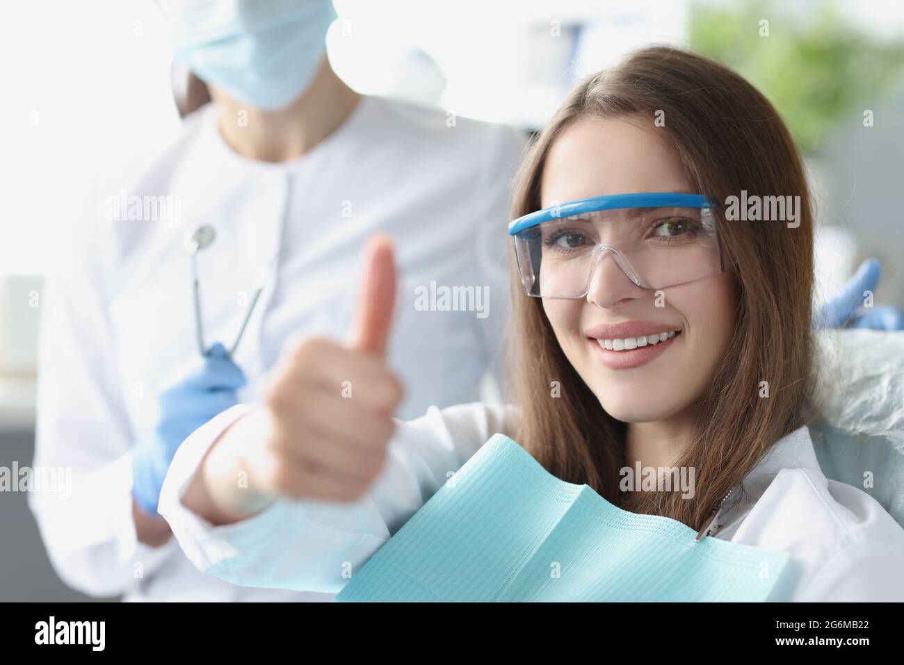 Junge Frau in einer Sicherheitsbrille, die auf einem Zahnarztstuhl sitzt und den Daumen nach oben zeigt Stockfoto