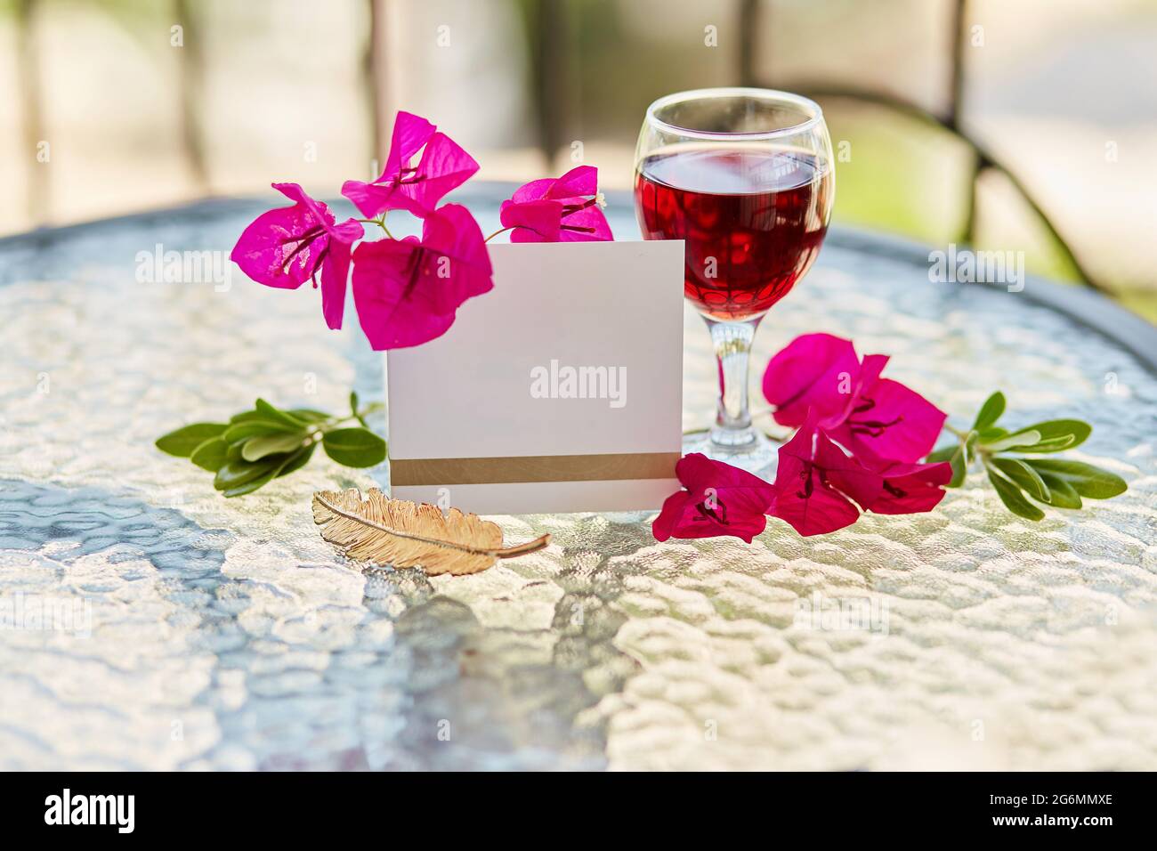 Ein Glas Rotwein auf dem Tisch auf der Terrasse. Dekorative rosa Blüten von Bougainvillea und goldenen Federzeichen. Nachbau einer Postkarte oder Einladung. Ro Stockfoto