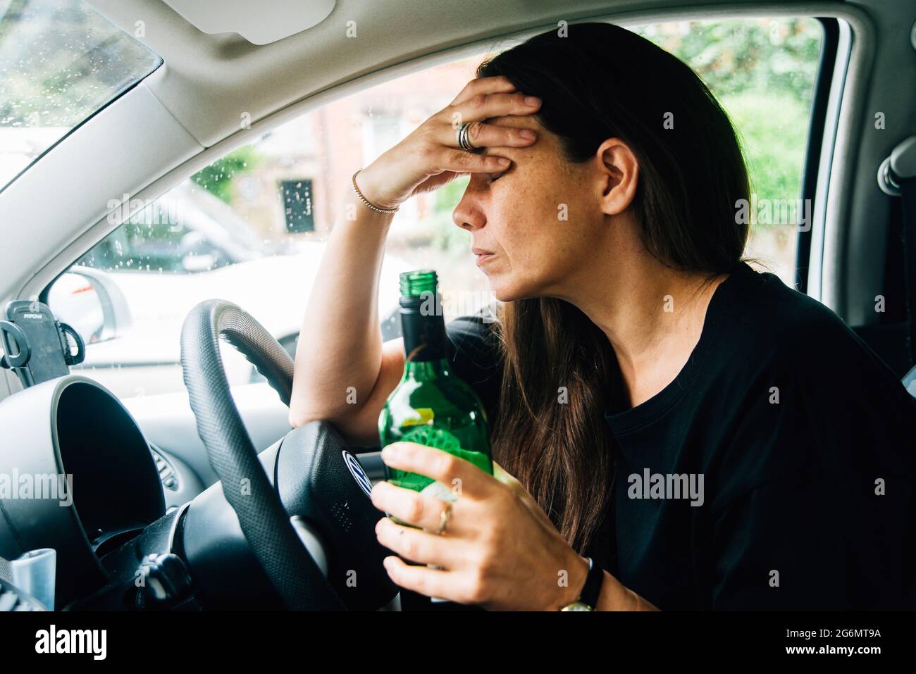 Eine Frau mit gemischter Rasse, die in ihrem Auto sitzt und eine Flasche Wein trinkt, wobei ihr Kopf in den Händen gestresst aussieht. Mutter mit Depression oder Angst Stockfoto