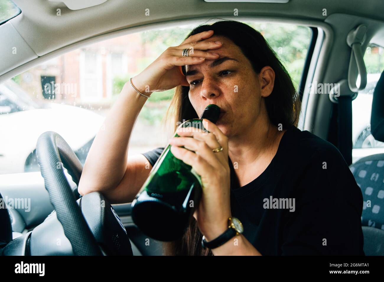 Eine Frau mit gemischter Rasse, die in ihrem Auto sitzt und eine Flasche Wein trinkt, wobei ihr Kopf in den Händen gestresst aussieht Stockfoto