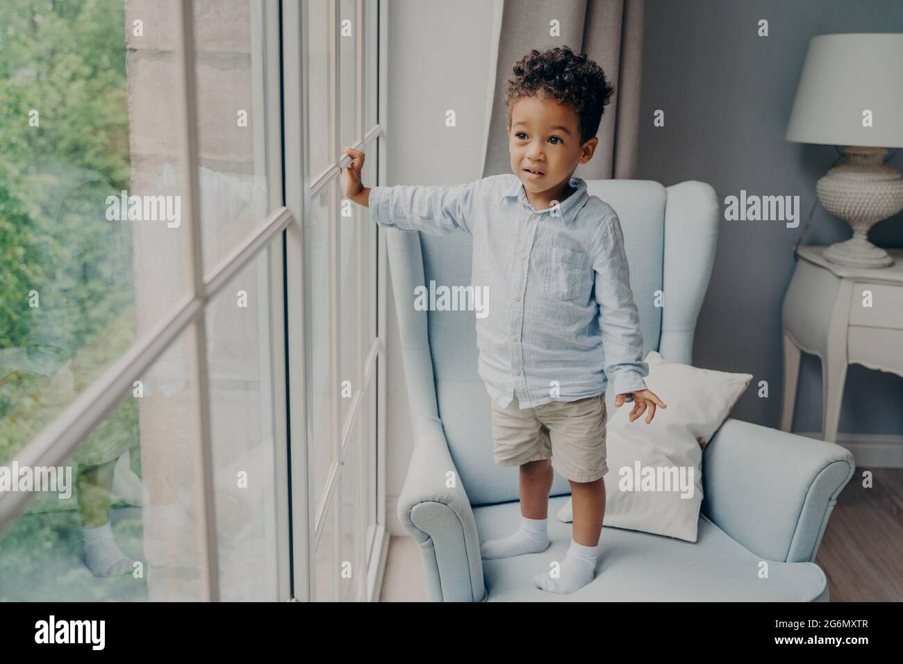 Neugieriger kleiner afroamerikanischer Junge, der durch das Fenster schaut, während er zu Hause auf einem Sessel steht Stockfoto