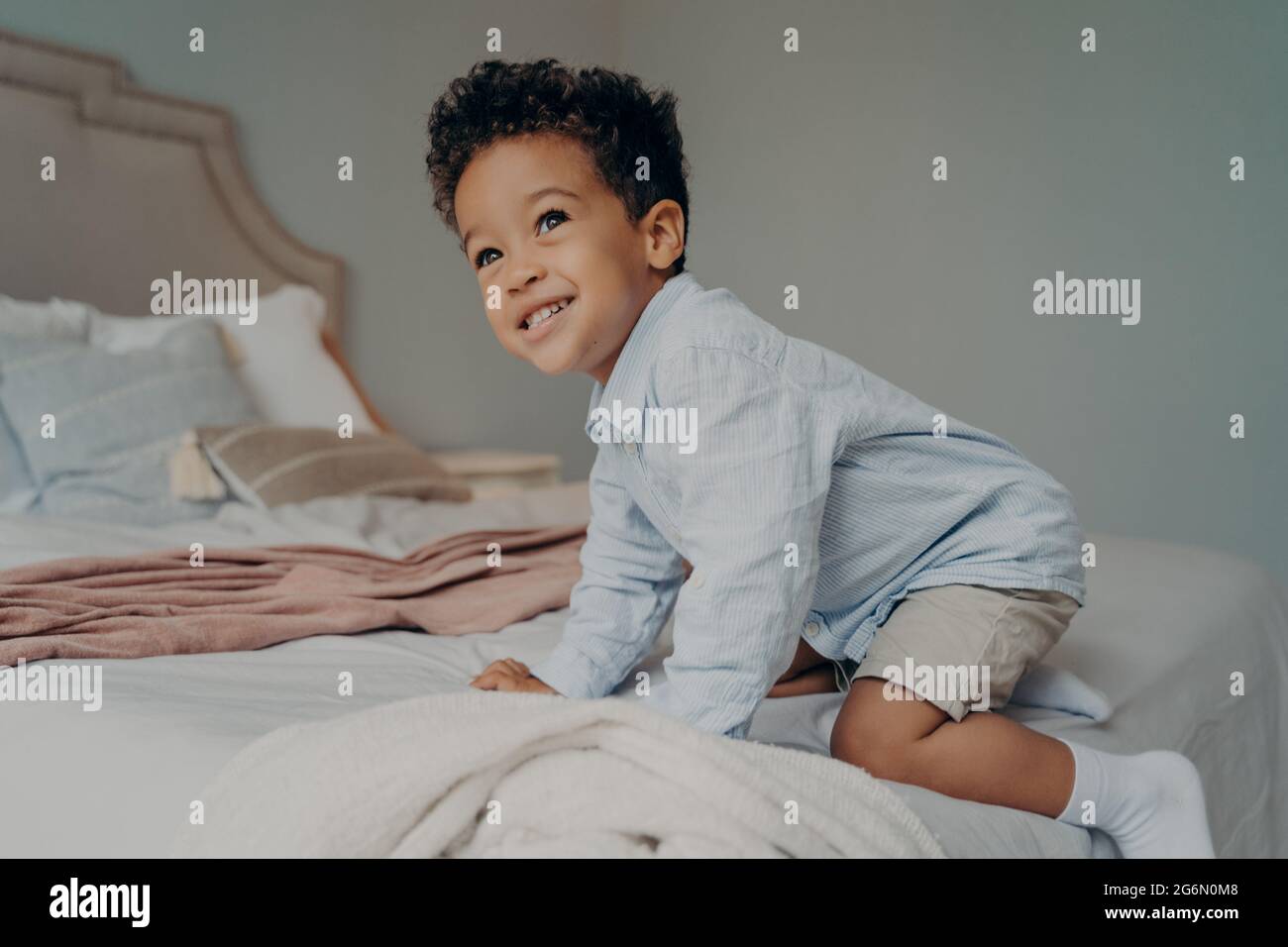 Lächelndes afroamerikanisches Kind, das zu Hause versucht, auf ein großes Bett zu klettern und sich drinnen amüsieren will Stockfoto