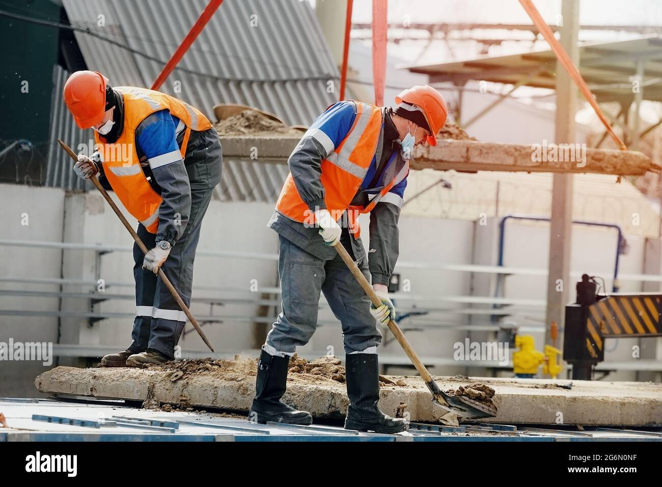 Zwei Arbeiter in Schutzmützen, Arbeitskleidung und einer medizinischen Maske arbeiten auf einer Baustelle mit Schaufeln. Harte körperliche Arbeit. Eine authentische Szene bei der Arbeit Stockfoto